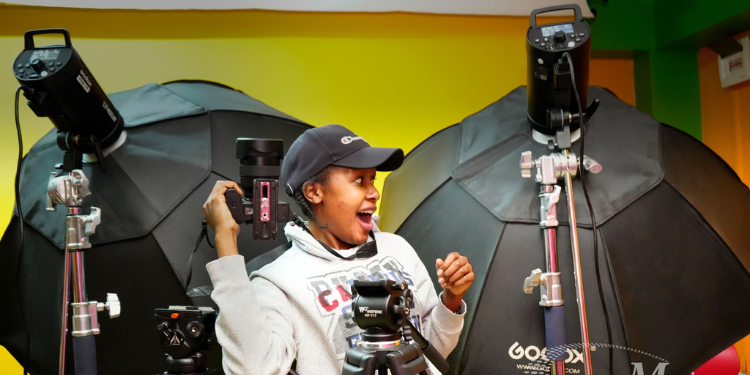 A Female Photography Student At Music And Film Academy In Nairobi Smiling While Holding A Professional Dslr Camera In A Studio Equipped With Godox Softboxes And Heavy-Duty Tripods.